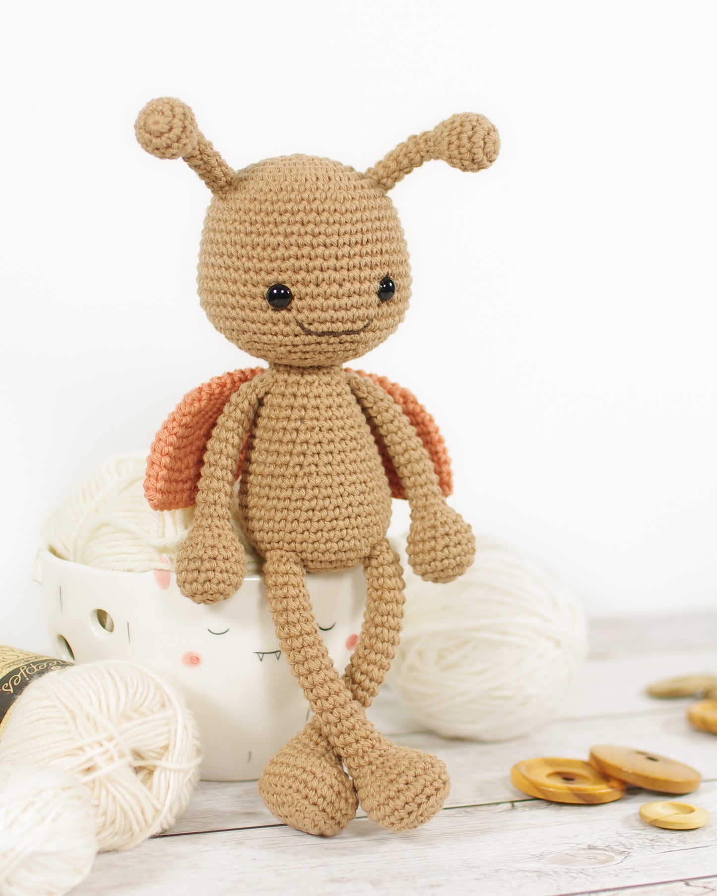 Crocheted toy ladybug with a light brown body and terracotta orange wings leaning against a yarn bowl.