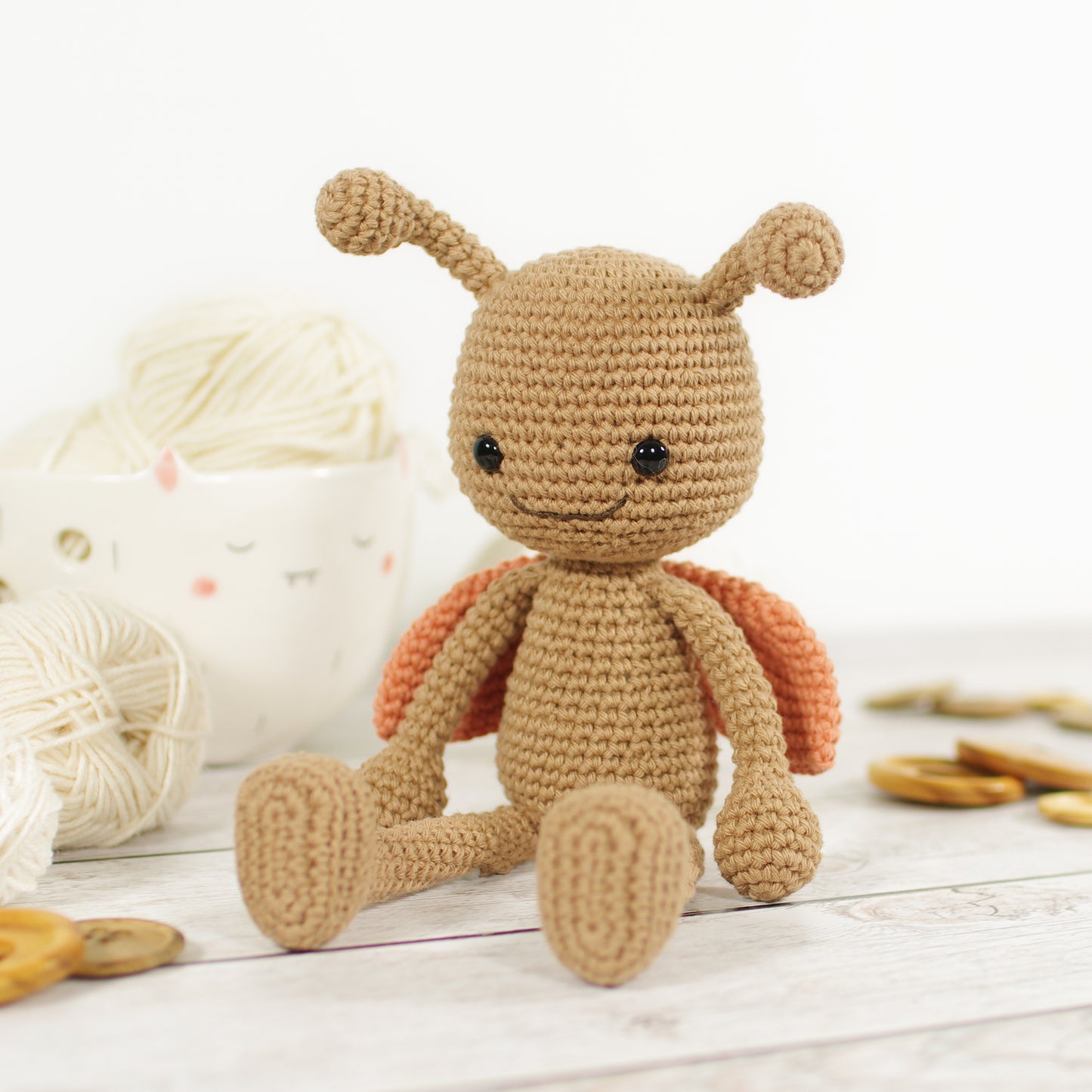 Crocheted long-legged toy ladybug sitting in front of yarn balls, ceramic yarn bowl and wooden buttons.