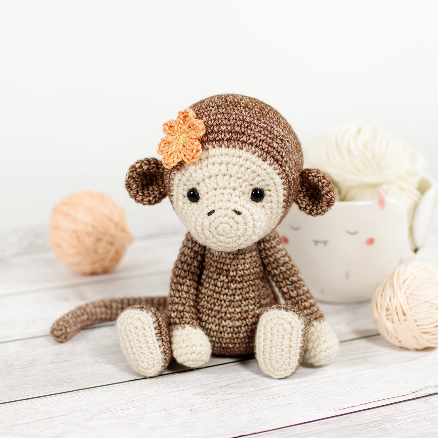 Brown crocheted monkey with cream face and peach flower on the head, sitting in front of balls of yarn.